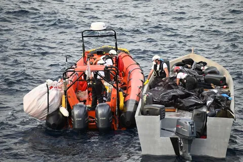 En gummibåt ligger intill en mindre träbåt på centrala Medelhavet. På träbåten ligger flera svarta säckar som två män hjälps åt att lyfta över till gummibåten.