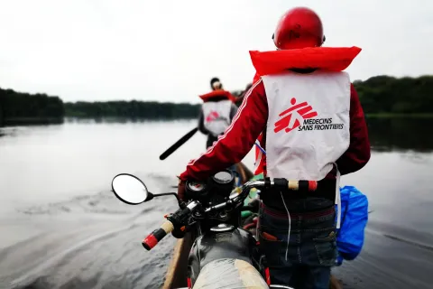 Två personer transporterar motorcyklar i en kanot över floden.