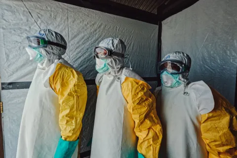 Tre personer i heltäckande eboladräkter står uppradade.